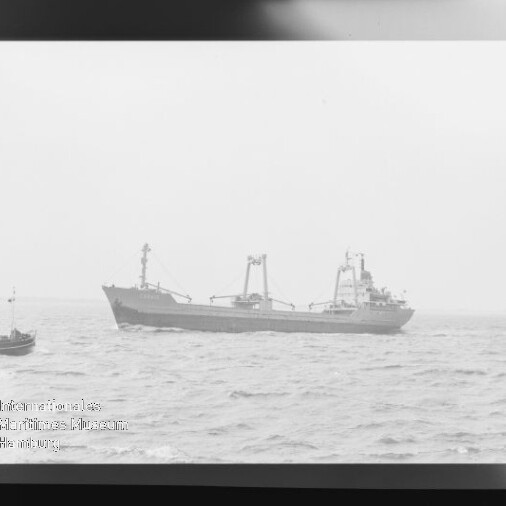 Schwarzweißaufnahme eines Frachtschiffs auf leichter See; links ein kleines Boot sowie sichtbarer Filmnegativrand und Museumslogo unten links.