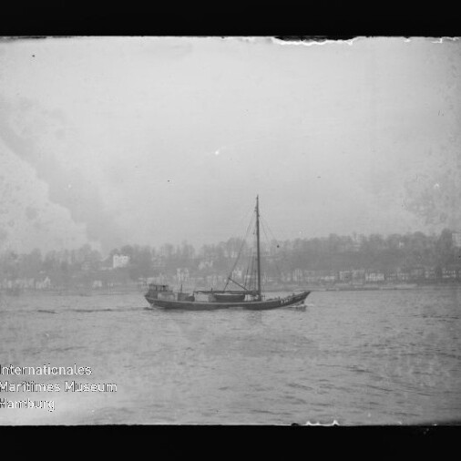 Schwarzweißfoto einer einmastigen Segelyacht auf ruhigem Wasser vor einer Uferlinie mit Häusern und Bäumen; am unteren linken Bildrand sichtbares Logo des Internationalen Maritimen Museums Hamburg.