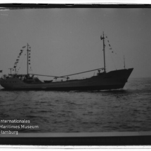 Schwarzweißaufnahme eines kleinen Handelsschiffs in Seitenansicht auf ruhiger See, mit zwei Masten, geschmückten Wimpelketten und dem Logo des Internationalen Maritimen Museums Hamburg unten links.