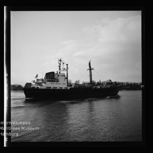 Schwarzweiß-Foto eines mittleren Frachtschiffs mit Kommandobrücke, Schornstein und zwei Masten, das auf ruhigem Wasser vor flachem Ufer fährt; quadratischer Negativrahmen und Logo des Internationalen Maritimen Museums Hamburg unten links sichtbar.