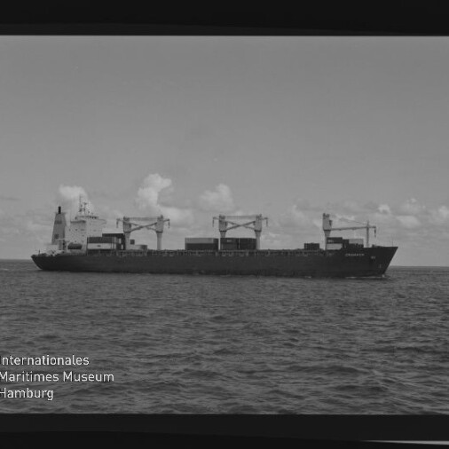 Schwarzweiß-Foto eines Frachtschiffs in ruhiger See, seitliche Ansicht mit drei sichtbaren Ladekränen und Containern auf dem Deck, flacher Horizont und vereinzelte Wolken im Hintergrund sowie Logo des Internationalen Maritimen Museums Hamburg in der linken unteren Ecke.