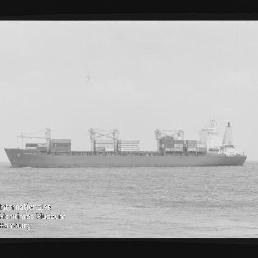 Schwarzweiß-Seitenansicht eines Containerschiffs mit aufgestapelten Containern und drei großen Deckskranen auf ruhiger See, mit schwarzem Filmrand und Museumslogo unten links