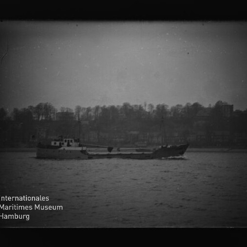 Schwarzweiß-Foto eines niedrigen Frachtschiffs, das auf ruhigem Wasser vor einer baumbestandenen Uferlinie mit Gebäudesilhouetten fährt; leicht körnig, mit Museumslogo unten links.