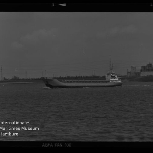 Schwarzweißaufnahme eines Frachtschiffs, das auf ruhigem Wasser an einem Industriehafen mit Fabrikgebäuden und Strommasten im Hintergrund vorbeifährt; links unten Museum-Logo und Filmrand mit der Aufschrift AGFA PAN 100.