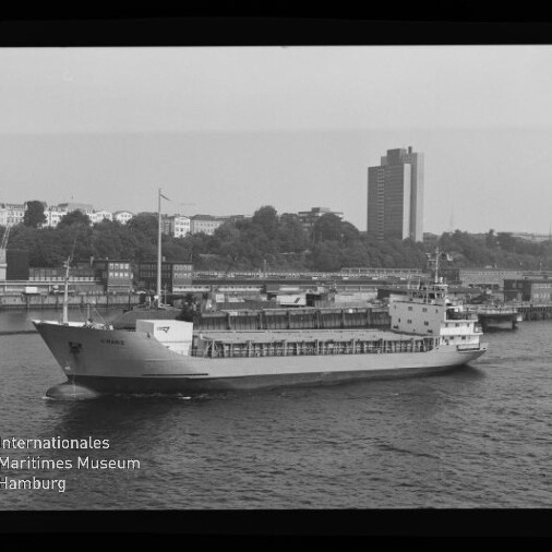 Schwarz-weiß-Foto eines Frachtschiffs, das am Kai eines Hafens vorbeifährt; im Hintergrund Uferbebauung mit Bäumen und einem hohen Büroturm, unten links das Wasserzeichen des Internationalen Maritimen Museums Hamburg.