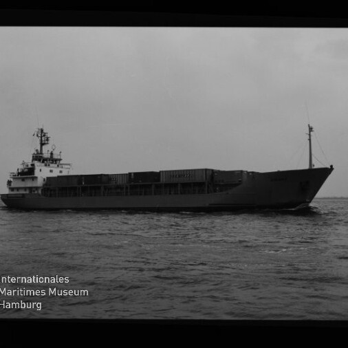 Schwarzweiß-Seitenansicht eines mittleren Frachtschiffs mit aufgestapelten Ladecontainern und sichtbarer Aufbauten auf ruhiger See; unten links Logo und Schriftzug des Internationalen Maritimen Museums Hamburg.