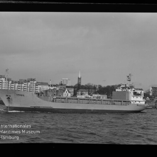 Schwarzweiß-Foto eines Frachtschiffs, das auf dem Wasser vor einer städtischen Uferfront mit markantem Kirchturm fährt; unten links Logo des Internationalen Maritimen Museums Hamburg und sichtbarer Filmrand mit der Ziffer 13.