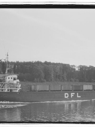 Schwarzweißaufnahme eines Frachtschiffs mit großen Buchstaben DFL am Rumpf, beladen mit Containern und fahrend auf einem ruhigen Kanal vor einem bewaldeten Ufer, mit Bug- und Heckwellen.