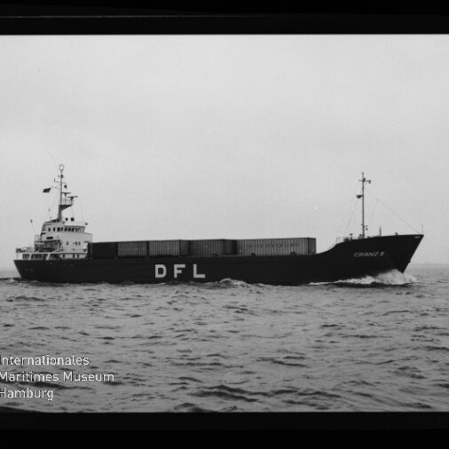 Schwarzweiß-Seitenaufnahme eines Frachtschiffs mit aufgestapelten Containern und großer weißer Aufschrift „DFL“ am Rumpf, das bei leicht bewegter See unter grauem Himmel fährt; Museumslogo unten links sichtbar.