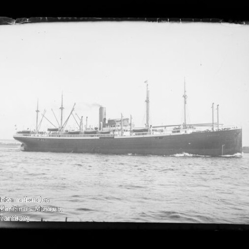 Seitliche Schwarzweißaufnahme eines großen Frachtdampfers mit mehreren Ladebäumen und einem zentralen Schornstein, der auf ruhiger See entlangfährt; unten links sichtbar das Wasserzeichen des Internationalen Maritimen Museums Hamburg.