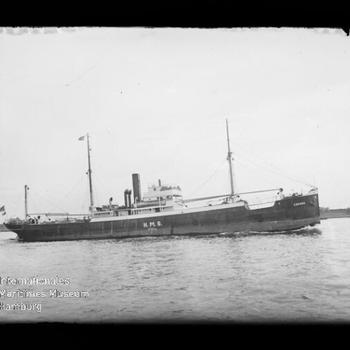 Schwarzweißaufnahme eines Frachtdampfers mit zwei Masten und einem Schornstein, die Aufschrift H.M.G. am Rumpf sichtbar, fahrend auf ruhigem Wasser vor flachem Horizont; Museumslogo unten links.