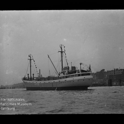 Schwarz-Weiß-Foto eines mittelgroßen Schiffes mit hellem Rumpf, zwei Masten und sichtbarer Takelage, vor einer Hafenpromenade mit Gebäuden im Hintergrund und Museumslogo unten links.