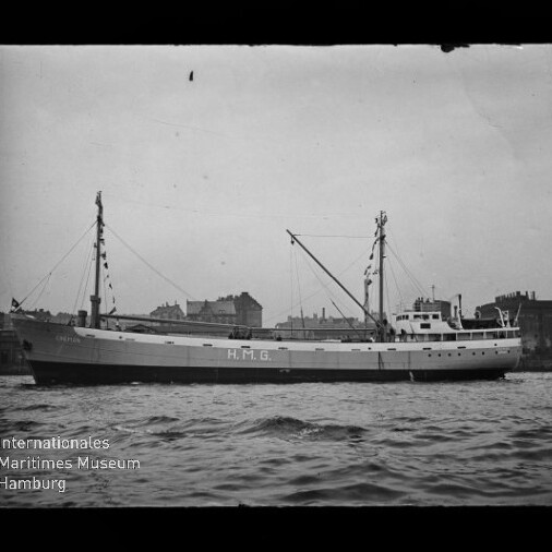 Schwarzweißaufnahme eines Frachtschiffs mit der großen Aufschrift H.M.G. am Rumpf, zwei Masten und Ladebäumen, Brücke achtern und einer Hafen-Silhouette im Hintergrund über welliger See.
