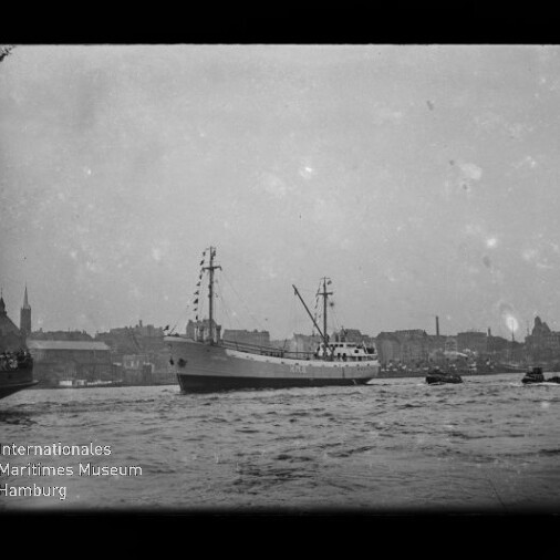 Schwarzweißaufnahme eines geschmückten Frachtschiffs mit zwei Masten, das im Hafen vor einer Stadtkulisse mit Kirchtürmen fährt; im Vordergrund unruhiges Wasser und kleine Begleitboote, links unten das Logo des Internationalen Maritimen Museums Hamburg.