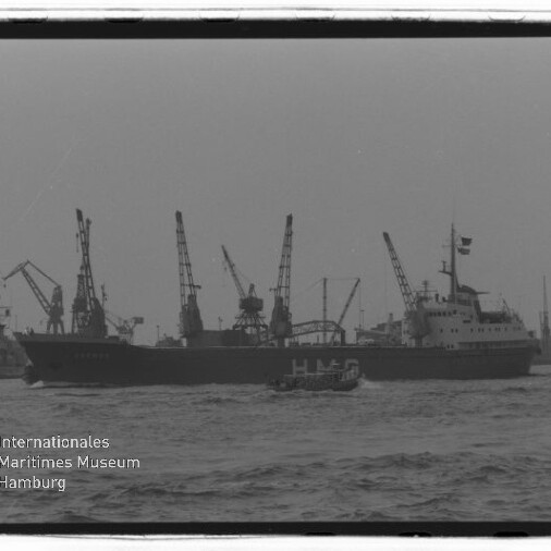 Schwarzweißfoto eines Frachtschiffs mit großen Buchstaben HMG auf der Bordwand, das in einem Industriehafen an mehreren Kränen vorbeifährt; im Vordergrund ein kleines Lotsenboot.