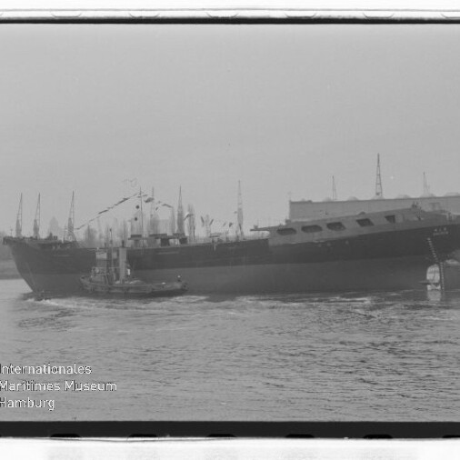 Schwarzweißaufnahme eines großen Frachtschiffs, das auf dem Wasser längsseits von einem Schlepper begleitet wird; im Hintergrund sind Hafenkräne und Lagerhallen zu sehen, unten links das Logo des Internationalen Maritimen Museums Hamburg.
