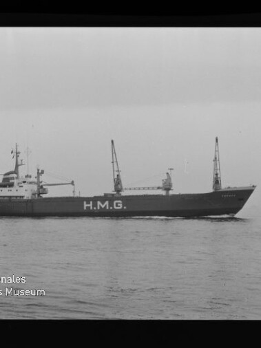 Schwarzweiß-Seitenansicht eines Frachtschiffs mit großer Aufschrift H.M.G., zwei Ladekränen auf dem Deck, ruhige See, sichtbarer Filmrand mit Markierungen und Museumslogo unten links.