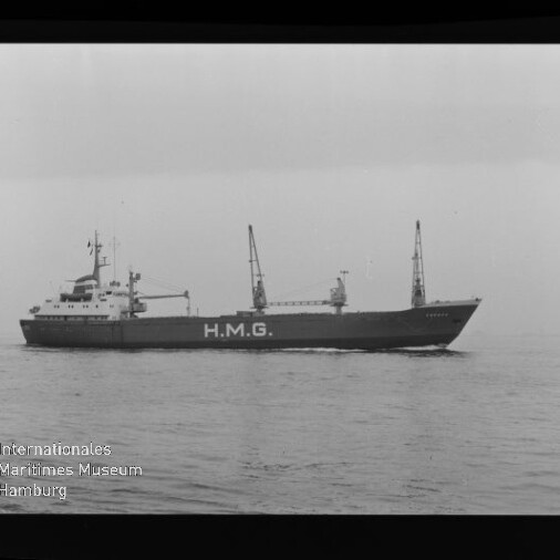 Schwarzweiß-Seitenansicht eines Frachtschiffs mit großer Aufschrift H.M.G., zwei Ladekränen auf dem Deck, ruhige See, sichtbarer Filmrand mit Markierungen und Museumslogo unten links.
