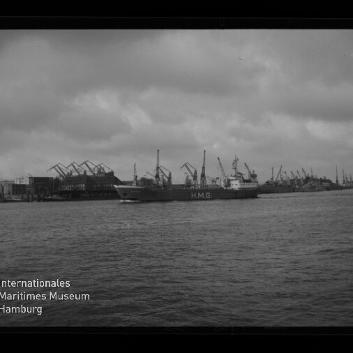 Schwarzweiß-Foto eines Frachtschiffs mit der Aufschrift H.M.G. auf dem Wasser vor einem Industriehafen mit zahlreichen Hafenkränen und einem bedeckten Himmel, unten links das Logo des Internationalen Maritimen Museums Hamburg