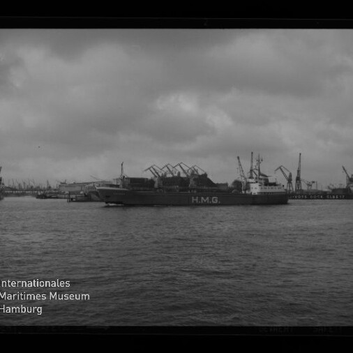 Frachtschiff mit großer Aufschrift H.M.G. vor einem Industriehafen mit zahlreichen Kränen und Lagerhallen, ruhige Wasserfläche im Vordergrund und bewölkter Himmel; Museumslogo unten links sichtbar.