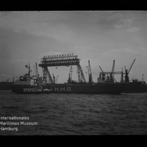 Schwarzweiß-Foto eines Frachtschiffs mit dem Schriftzug H.M.G. vor einer Werft, über der ein großer Gitterkran mit dem Schriftzug „Stülckenwerft“ steht; ein Schlepper an der Seite, Wasser im Vordergrund und das Logo des Internationalen Maritimen Museums Hamburg unten links.