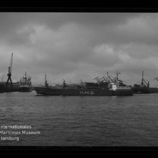 Frachtschiff mit der Aufschrift H.M.G. fährt in einem Industriehafen vor einer Reihe von Kränen unter einem bewölkten Himmel; im Vordergrund Wasser und unten links das Logo des Internationalen Maritimen Museums Hamburg.