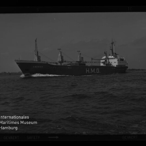 Schwarzweiß-Foto eines Frachtschiffs mit großer Aufschrift „H.M.G.“ am Rumpf und mehreren Ladekränen, das mit Bugwelle über die See fährt; im Hintergrund flacher Küstenstreifen und ein kleines Segelboot, links unten Museumswasserzeichen.