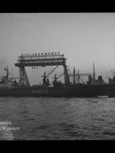 Schwarzweißaufnahme eines Frachtschiffs mit großer Aufschrift H.M.G. im Hafen vor einem hohen Werftkran mit dem Schriftzug STULCKENWERFT; mehrere Ladekräne und weitere Schiffe im Hintergrund, im Vordergrund Wasser und unten links das Logo des Internationalen Maritimen Museums Hamburg.