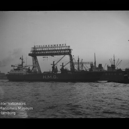 Schwarzweißaufnahme eines Frachtschiffs mit großer Aufschrift H.M.G. im Hafen vor einem hohen Werftkran mit dem Schriftzug STULCKENWERFT; mehrere Ladekräne und weitere Schiffe im Hintergrund, im Vordergrund Wasser und unten links das Logo des Internationalen Maritimen Museums Hamburg.