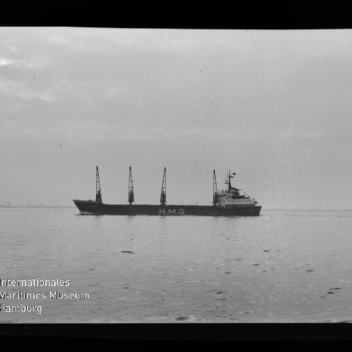 Schwarzweißaufnahme eines Frachtschiffs mit der Aufschrift H.M.G. und mehreren Ladekränen, das ruhig über spiegelglattes Wasser vor einem flachen Horizont fährt.