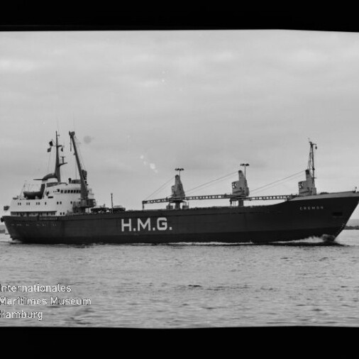 Schwarzweißfoto eines Frachtschiffs mit großer Aufschrift H.M.G. am Rumpf, drei Ladekräne und das Deckshaus achtern sichtbar, das auf ruhiger See fährt; links unten Wasserzeichen des Internationalen Maritimen Museums Hamburg.
