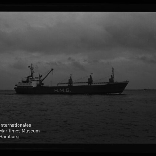 Schwarzweiß-Foto eines Frachtschiffs mit der Aufschrift H.M.G. auf dem Rumpf, das bei bedecktem Himmel auf ruhiger See vorbeifährt; links unten das Logo des Internationalen Maritimen Museums Hamburg und am rechten Bildrand der Filmrand mit der Aufschrift AGFA LISS sichtbar.