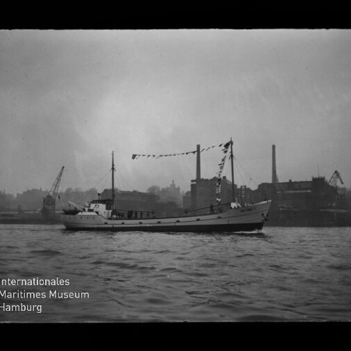 Schwarzweißaufnahme eines geschmückten Küsten- oder Arbeitsschiffs mit Flaggenbändern, das auf ruhigem Wasser vor einer industriellen Hafenfront mit Kränen und Fabrikschornsteinen fährt; Logowasserzeichen des Internationalen Maritimen Museums Hamburg in der linken unteren Ecke.