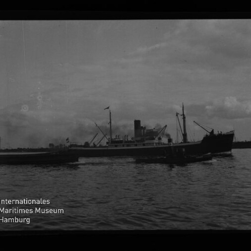 Schwarzweiß-Foto eines größeren Frachtdampfers mit markantem Schornstein und Ladekränen, neben einem kleineren Begleitboot auf ruhiger See unter bewölktem Himmel; unten links das Logo und Schriftzug des Internationalen Maritimen Museums Hamburg.