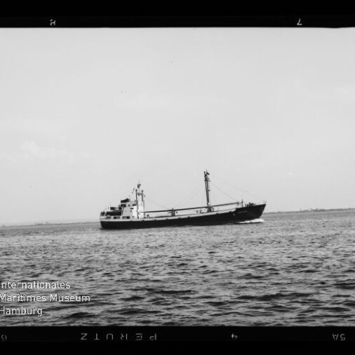 Schwarzweißaufnahme eines Frachtschiffs auf ruhiger See, seitliche Ansicht mit niedrigem Deckshaus am Heck und mittschifflichem Ladebaum/Mast vor flachem Horizont; Wasserzeichen des Internationalen Maritimen Museums Hamburg links unten.