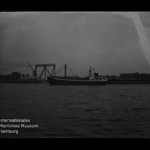 Frachtschiff auf ruhigem Wasser vor der Silhouette eines Industriehafens mit Kränen und einer großen Hubbrücke, dunkle kontrastarme Aufnahme mit Museumslogo unten links.