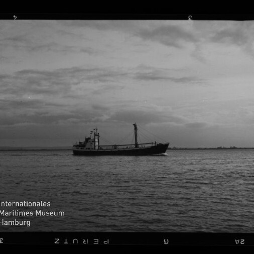 Schwarzweißaufnahme eines einzelnen Frachtschiffs in mittlerer Entfernung auf ruhiger See vor einer bewölkten Horizontlinie; Filmrand und Logo des Internationalen Maritimen Museums Hamburg unten links sichtbar.
