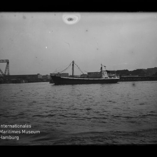 Schwarzweiß-Foto eines Frachtschiffs mit geschmückten Signalflaggen, seitlich auf der Wasseroberfläche vor einem Kai mit Industrieanlagen und einer Hubbrücke; unten links das Wasserzeichen des Internationalen Maritimen Museums Hamburg.