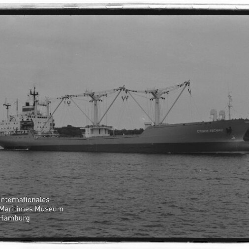 Schwarzweißfoto eines großen Frachtschiffs in Seitenansicht mit mehreren Ladekränen, das auf ruhigem Wasser fährt; im Vordergrund rechts ein kleines Segelboot und links unten das Logo des Internationalen Maritimen Museums Hamburg.