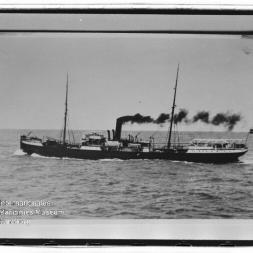 Schwarzweiß-Foto eines seitlich fahrenden Dampfschiffs mit mittigem Schornstein, der schwarzen Rauch ausstößt, zwei Masten, Flagge am Heck und leicht bewegter See, mit weißem Bildrand und Museumslogo unten links.