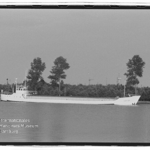 Schwarz-weiß-Foto eines langen, niedrigen Frachtschiffs mit weißem Rumpf, das ruhig auf dem Wasser vor einer Uferlinie mit einer Reihe hoher Bäume fährt; unten links ist ein Museumswasserzeichen sichtbar.