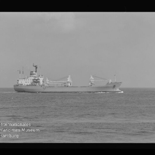 Schwarzweißaufnahme eines Frachtschiffs in Fahrt auf ruhiger See, seitlich zu sehen mit zwei Ladekränen und mittig liegenden Aufbauten vor dem Horizont; links unten ist ein Wasserzeichen des Internationalen Maritimen Museums Hamburg und ein schwarzer Filmrand erkennbar.
