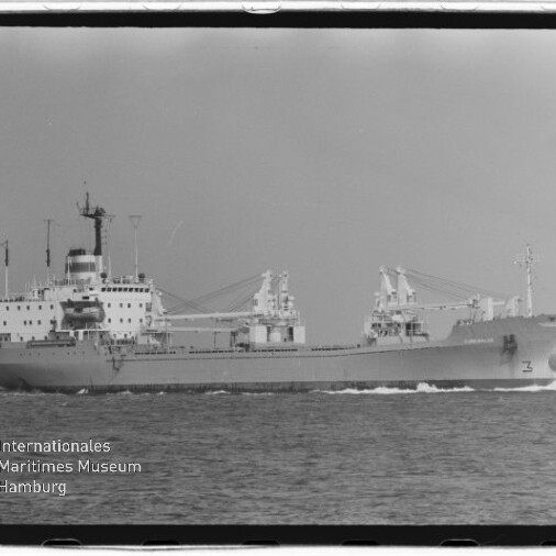Schwarzweiß-Foto eines Frachtschiffs in Seitenansicht mit Aufbauten achtern und mehreren Deckskränen, das nach rechts fahrend auf ruhiger See eine Bugwelle zieht; am unteren linken Bildrand Wasserzeichen des Internationalen Maritimen Museums Hamburg.