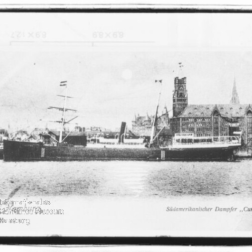 Schwarzweiß-Aufnahme eines südamerikanischen Dampfers namens „Curityba“ mit zwei Masten und Schornstein, liegend an einem Kai vor einem großen historischen Backsteingebäude mit Turm und spitzer Kirchturm-Silhouette, links unten ein rundes Wasserzeichen.