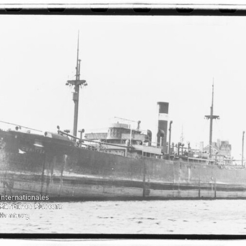 Schwarzweißes Foto eines großen Dampffrachters mit hohem Schornstein und mehreren Masten, seitlich im Hafen festgemacht, Wasser im Vordergrund und Gebäude im Hintergrund, unten links Wasserzeichen des Internationalen Maritimen Museums Hamburg.