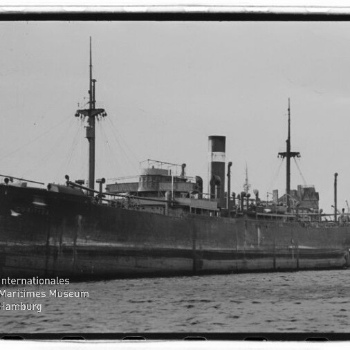 Schwarzweißaufnahme eines großen, am Kai festgemachten Dampffrachters mit hohem Schornstein und zwei Masten, im Vordergrund Wasser und unten links das Logo des Internationalen Maritimen Museums Hamburg.