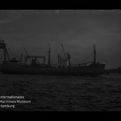 Schwarzweißaufnahme eines Frachtschiffs im Hafen vor Krananlagen und Pier, mit welliger See im Vordergrund und dem Logo des Internationalen Maritimen Museums Hamburg in der linken unteren Ecke.