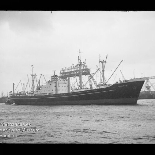Schwarzweißaufnahme eines großen Frachtschiffs mit Ladebäumen und mehreren Masten, seitlich im Hafen auf ruhigem Wasser liegend, im Hintergrund Hafenkräne und Industrieanlagen.
