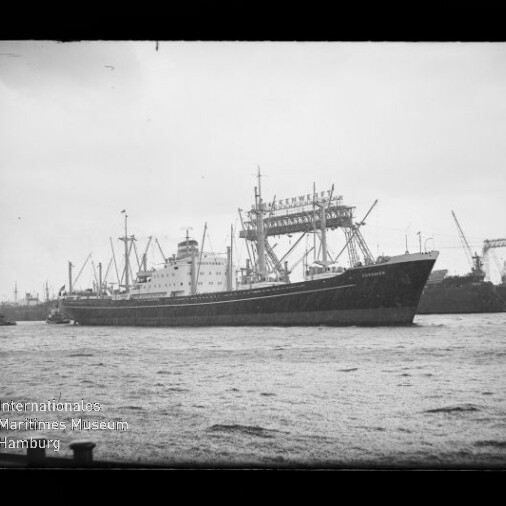 Schwarzweiß-Foto eines großen Frachtschiffs mit mehreren Ladekränen und sichtbaren Aufbauten, das in einem Industriehafen mit hohen Portalkränen im Hintergrund fährt; im Vordergrund Wasser und unten links das Logo des Internationalen Maritimen Museums Hamburg.