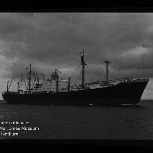 Schwarzweiß-Foto eines großen Frachtschiffs mit mehreren Ladekränen und sichtbaren Aufbauten, das bei bewölktem Himmel auf ruhigem Meer fährt; Filmrand mit Beschriftung ‚ADOX R 21‘ und Logo des Internationalen Maritimen Museums Hamburg im Bildrand.
